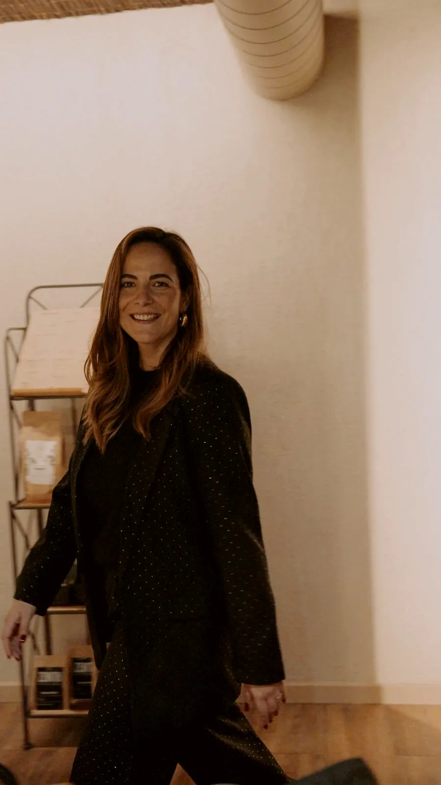Mulher de blazer escuro a atravessar uma sala com luz quente, a sorrir.