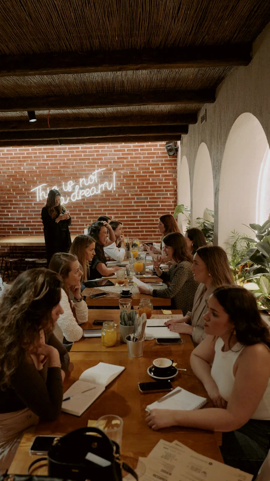 Mesa longa num espaço de tijolo e arcos, mulheres em conversa, cadernos abertos e chávenas de café.